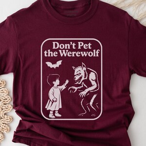 May include: Burgundy t-shirt with a white graphic. The graphic features the text "Don't Pet the Werewolf" above an illustration of a child reaching toward a werewolf. A bat graphic is above the child and werewolf.