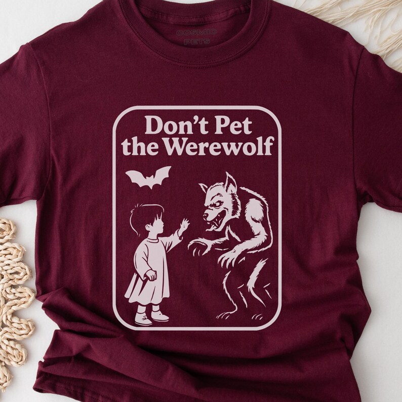May include: Burgundy t-shirt with a white graphic. The graphic features the text "Don't Pet the Werewolf" above an illustration of a child reaching toward a werewolf. A bat graphic is above the child and werewolf.