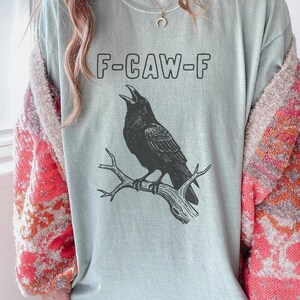 May include: A light blue t-shirt with the text "F-CAW-F" above a black raven perched on a branch. The raven is depicted with its beak open. The shirt is a casual style.