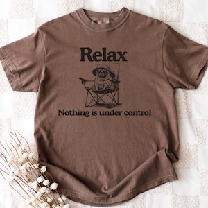 May include: A brown t-shirt with the words "Relax" and "Nothing is under control" printed in black. The shirt features a graphic of a pug wearing a hat and sunglasses, sitting in a folding chair.
