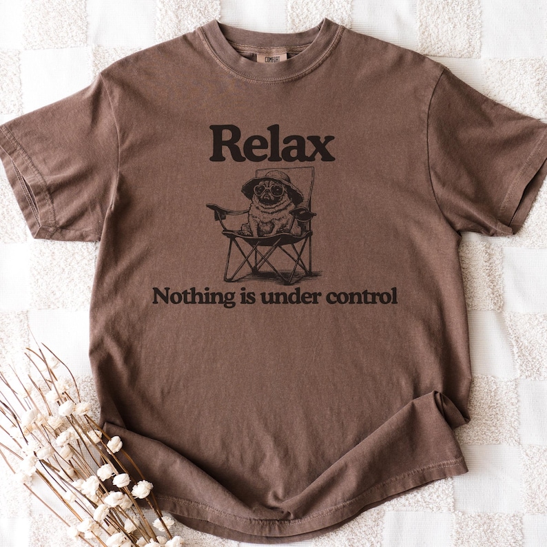 May include: A brown t-shirt with the words "Relax" and "Nothing is under control" printed in black. The shirt features a graphic of a pug wearing a hat and sunglasses, sitting in a folding chair.