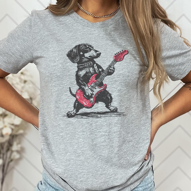K&ouml;nnte beinhalten: Graues T-Shirt mit einem schwarz-wei&szlig;en Dackel, der eine rote E-Gitarre spielt. Der Hund tr&auml;gt ein Halsband. Die Grafik ist eine detaillierte Illustration. Das Shirt ist aus einem weichen Material.