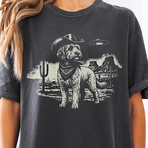 Puede incluir: Una camiseta gris carbón con un estampado gráfico de un perro con sombrero de vaquero y pañuelo. La escena incluye un paisaje desértico con un cactus saguaro, formaciones rocosas y un platillo volante flotando en el cielo sobre el perro.