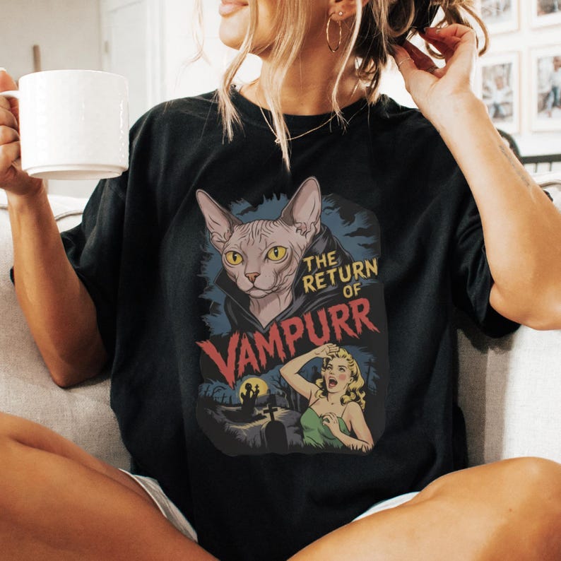 Peut inclure: T-shirt noir avec un motif d'un chat Sphynx aux yeux jaunes, portant une cape de vampire, et le texte "THE RETURN OF VAMPURR". Le design comprend une sc&egrave;ne de cimeti&egrave;re et une femme effray&eacute;e.