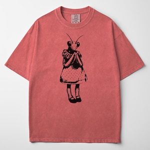 Puede incluir: Camiseta de color coral con una ilustraci&oacute;n negra de una mantis religiosa vestida con un vestido de lunares. El insecto se representa con las manos juntas. La camiseta tiene cuello redondo y mangas cortas.