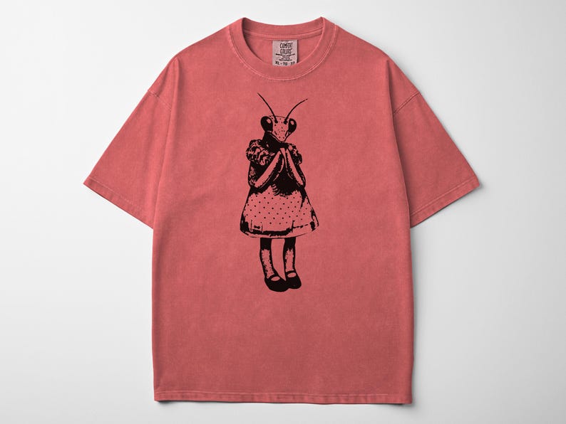 Puede incluir: Camiseta de color coral con una ilustraci&oacute;n negra de una mantis religiosa vestida con un vestido de lunares. El insecto se representa con las manos juntas. La camiseta tiene cuello redondo y mangas cortas.