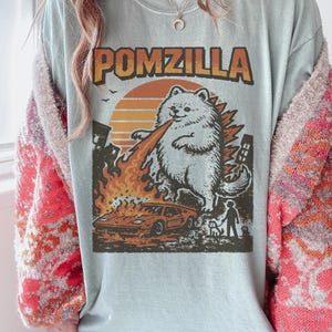 May include: A pale blue t-shirt featuring the text "POMZILLA" above a cartoon Pomeranian dog breathing fire. The dog is shown as a monster destroying a city, with a car and a person with a dog in the foreground.