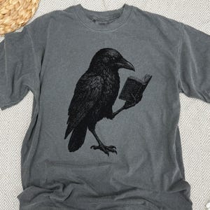 May include: Gray t-shirt featuring a black illustration of a raven reading a book. The raven is detailed with textured feathers and is holding the book with its clawed foot. The shirt has a crew neck and short sleeves.