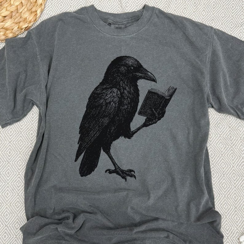 May include: Gray t-shirt featuring a black illustration of a raven reading a book. The raven is detailed with textured feathers and is holding the book with its clawed foot. The shirt has a crew neck and short sleeves.