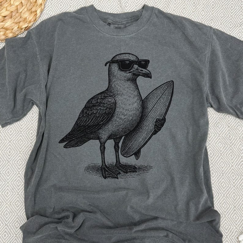 K&ouml;nnte beinhalten: Ein anthrazitfarbenes Kurzarm-T-Shirt mit einer schwarzen Grafik im Tusche-Stil, die eine M&ouml;we mit Sonnenbrille zeigt, die ein Surfbrett unter dem Fl&uuml;gel h&auml;lt. Das T-Shirt hat einen Rundhalsausschnitt und eine verwaschene Stoffstruktur.