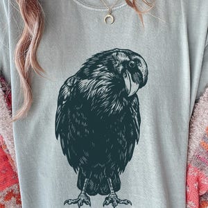 May include: A light blue t-shirt featuring a detailed black illustration of a raven. The raven is depicted in a detailed, artistic style, with its feathers and beak clearly defined. The shirt is made of a soft material.