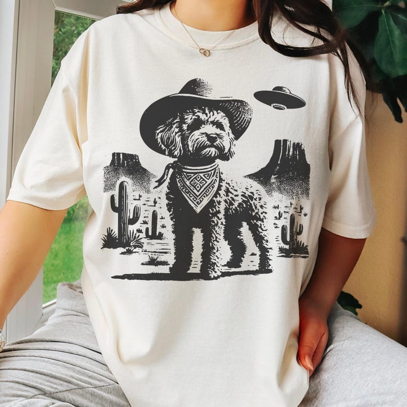 Pu&ograve; includere: T-shirt color crema con grafica in bianco e nero di un cane con cappello da cowboy e bandana, ambientato in un paesaggio desertico con cactus e montagne. Un UFO fluttua sopra il cane. La maglietta ha una vestibilit&agrave; ampia.