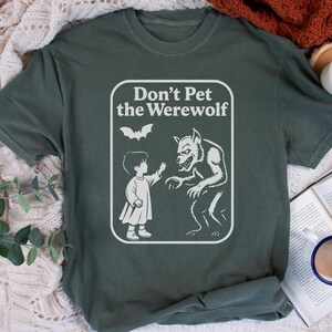 May include: Dark green t-shirt with a white graphic that reads "Don't Pet the Werewolf." The graphic features a child reaching toward a werewolf, with a bat above them. The shirt is made of a soft material.