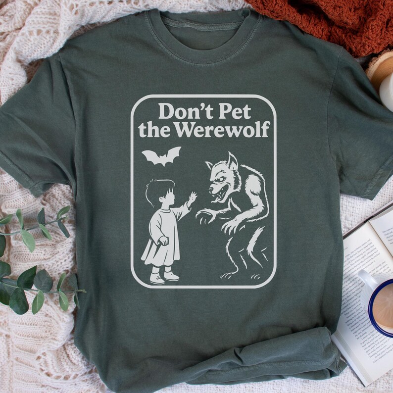 May include: Dark green t-shirt with a white graphic that reads "Don't Pet the Werewolf." The graphic features a child reaching toward a werewolf, with a bat above them. The shirt is made of a soft material.