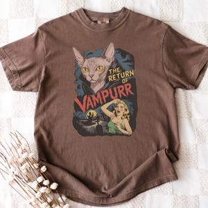 Peut inclure: T-shirt marron avec un motif d'un chat Sphynx en cape de vampire et le texte "THE RETURN OF VAMPURR". Le motif comprend &eacute;galement une femme hurlante et une sc&egrave;ne de cimeti&egrave;re.