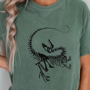 Op de afbeelding: Saliegroen t-shirt met een zwart dinosaurus skelet ontwerp. De afbeelding toont een gestileerd raptor skelet met een open mond en gebogen staart. Het shirt is een comfortabele, casual stijl.