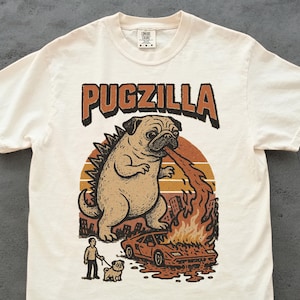 以下が含まれることがあります： 「PUGZILLA」の文字と、ゴジラ風のパグが描かれたクリーム色のTシャツ。パグは車に火を吹きかけ、人間と別のパグが見ている。デザインは茶色、オレンジ、黒の色合い。
