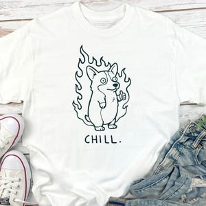 May include: White t-shirt featuring a black and white cartoon corgi dog with flames around it, giving a thumbs up. The word "CHILL." is printed below the dog. The shirt is paired with blue jeans and white sneakers.