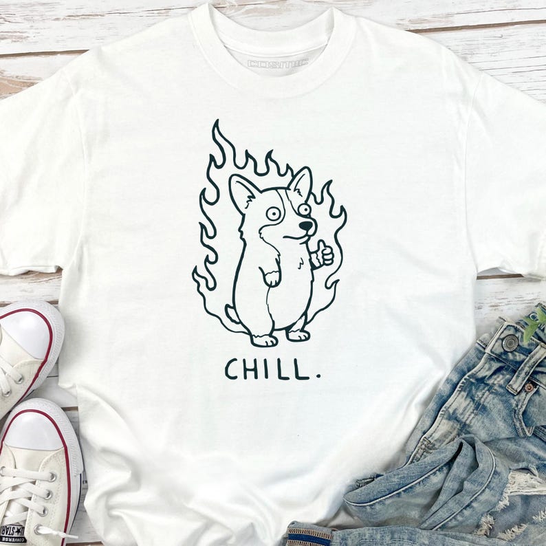 May include: White t-shirt featuring a black and white cartoon corgi dog with flames around it, giving a thumbs up. The word "CHILL." is printed below the dog. The shirt is paired with blue jeans and white sneakers.
