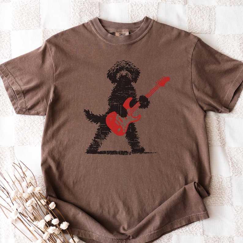 May include: Brown t-shirt featuring a black silhouette of a dog playing a red electric guitar. The dog is standing with its legs spread, holding the guitar. The shirt has a crew neck and short sleeves.