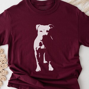 Pode incluir: Camiseta bord&ocirc; com uma silhueta branca de um cachorro. O cachorro &eacute; retratado em p&eacute;, com a cabe&ccedil;a ligeiramente virada. A camiseta tem gola redonda e mangas curtas. O texto "REBEL PETS" est&aacute; impresso no topo.
