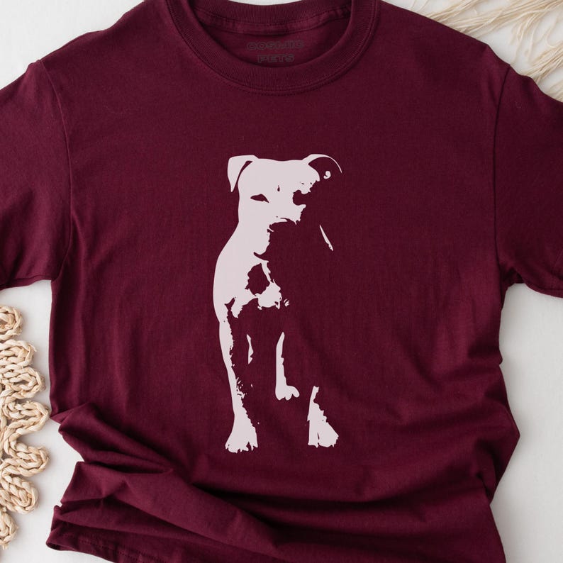 Pode incluir: Camiseta bord&ocirc; com uma silhueta branca de um cachorro. O cachorro &eacute; retratado em p&eacute;, com a cabe&ccedil;a ligeiramente virada. A camiseta tem gola redonda e mangas curtas. O texto "REBEL PETS" est&aacute; impresso no topo.
