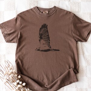 May include: A brown short-sleeved t-shirt featuring a black graphic of a bird in flight. The bird's wings are spread wide, showcasing detailed feather patterns. The shirt is made of a soft material and has a classic crew neck.