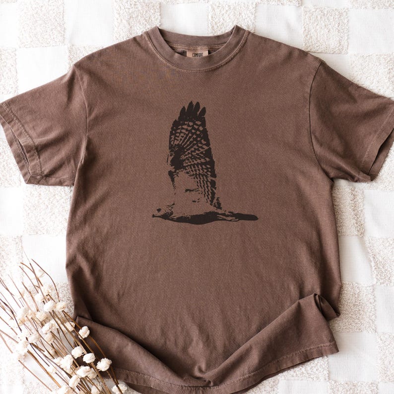 May include: A brown short-sleeved t-shirt featuring a black graphic of a bird in flight. The bird's wings are spread wide, showcasing detailed feather patterns. The shirt is made of a soft material and has a classic crew neck.