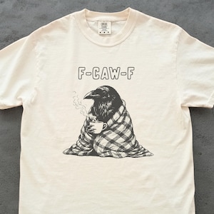 May include: Cream-colored t-shirt featuring a black and white illustration of a raven wrapped in a plaid blanket, holding a cup of coffee. The text "F-CAW-F" is printed above the raven. The shirt has short sleeves and a crew neck.