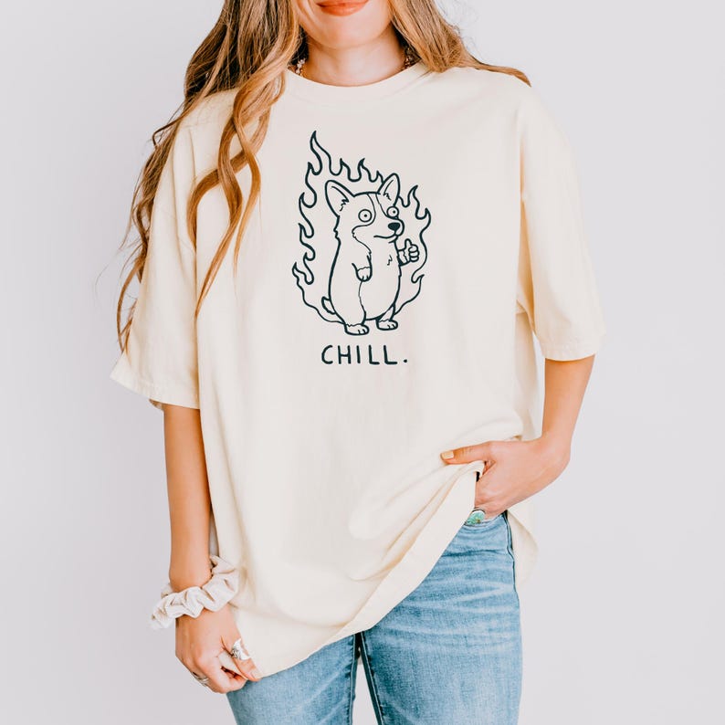 May include: Cream-colored t-shirt featuring a cartoon corgi dog in flames, giving a thumbs-up. The word "CHILL." is printed below the graphic in black. The shirt has a relaxed fit and short sleeves.