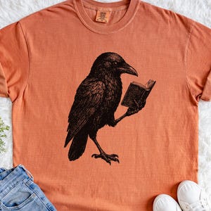 May include: Orange t-shirt featuring a black illustration of a raven reading a book. The raven is detailed with intricate feather patterns. The shirt has a crew neck and short sleeves. The shirt is made of a soft material.