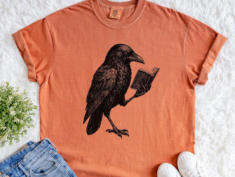 May include: Orange t-shirt featuring a black illustration of a raven reading a book. The raven is detailed with intricate feather patterns. The shirt has a crew neck and short sleeves. The shirt is made of a soft material.