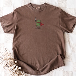 May include: Brown t-shirt featuring a cartoon frog wearing a red cape and holding a sword. The frog is green with a white sword and the word "Huzzah" is printed below the frog.