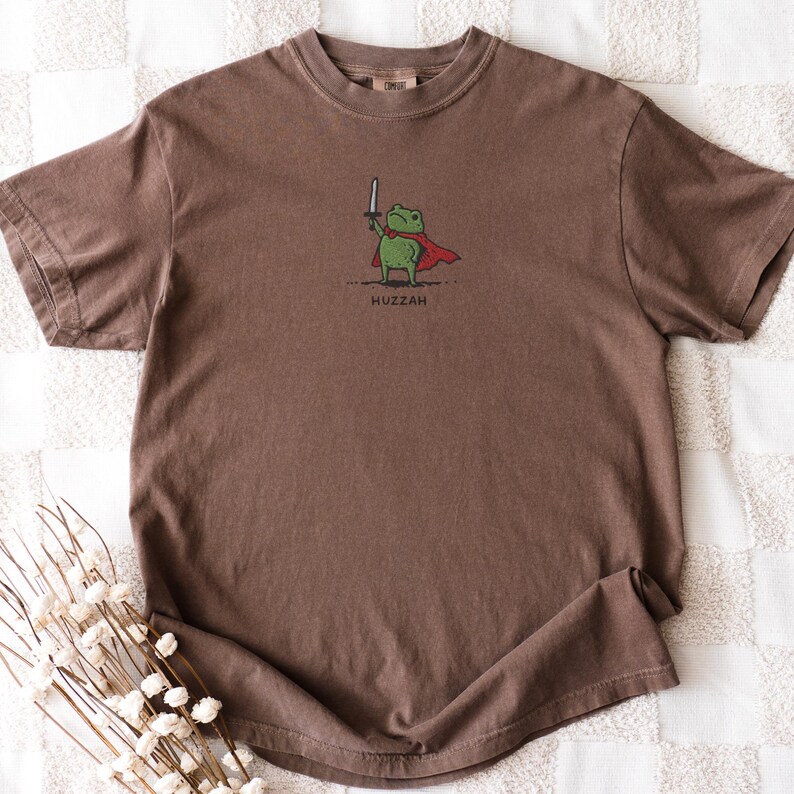 May include: Brown t-shirt featuring a cartoon frog wearing a red cape and holding a sword. The frog is green with a white sword and the word "Huzzah" is printed below the frog.