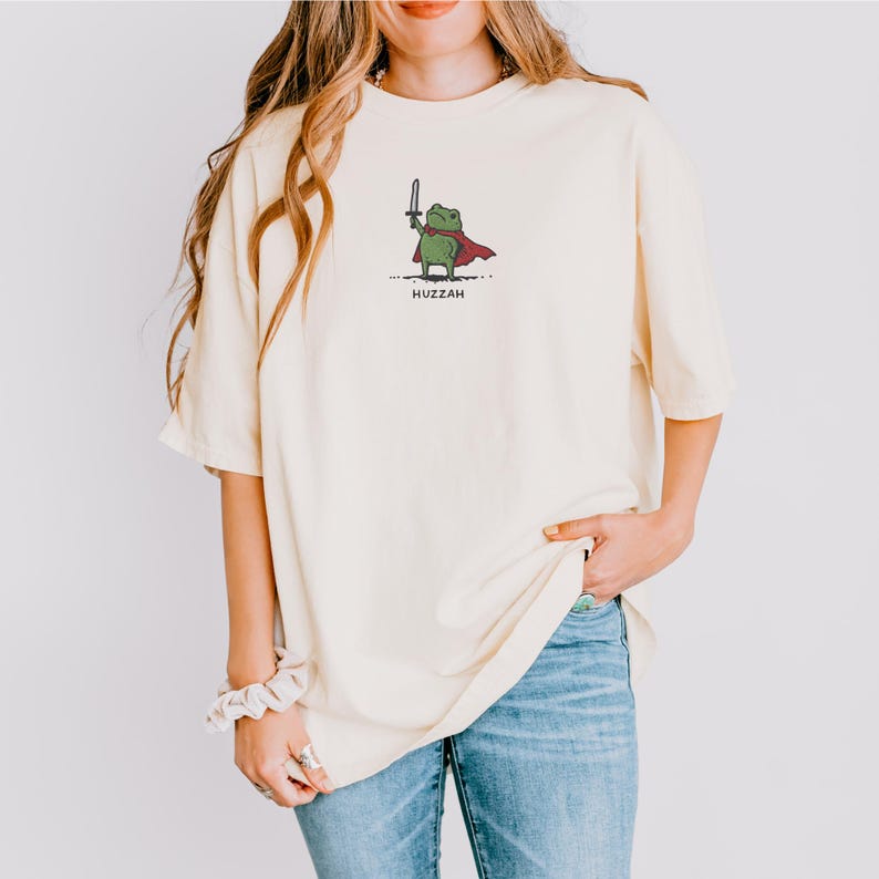 May include: Cream-colored t-shirt featuring a cartoon frog holding a sword and wearing a red cape. The word "Huzzah" is printed below the frog. The shirt is paired with blue jeans.
