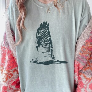 May include: A light green t-shirt featuring a dark green graphic of a bird in flight. The bird's wings are spread wide, showcasing detailed feather patterns. The shirt has a crew neck and short sleeves.