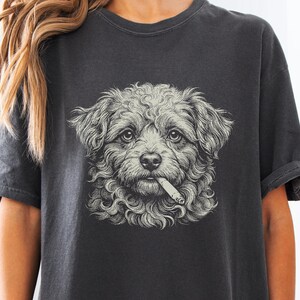 May include: A charcoal grey short-sleeved t-shirt featuring a detailed, light-coloured graphic illustration of a curly-haired dog with a cigarette in its mouth. The design is centred on the chest, showcasing intricate line work and shading.