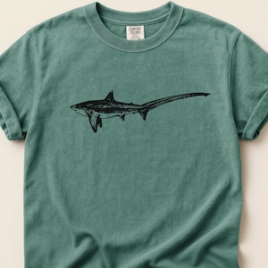 May include: A teal-colored t-shirt featuring a black illustration of a thresher shark. The shark is depicted in profile, with its distinctive long tail prominently displayed. The shirt has rolled-up sleeves and a Comfort Colors tag.