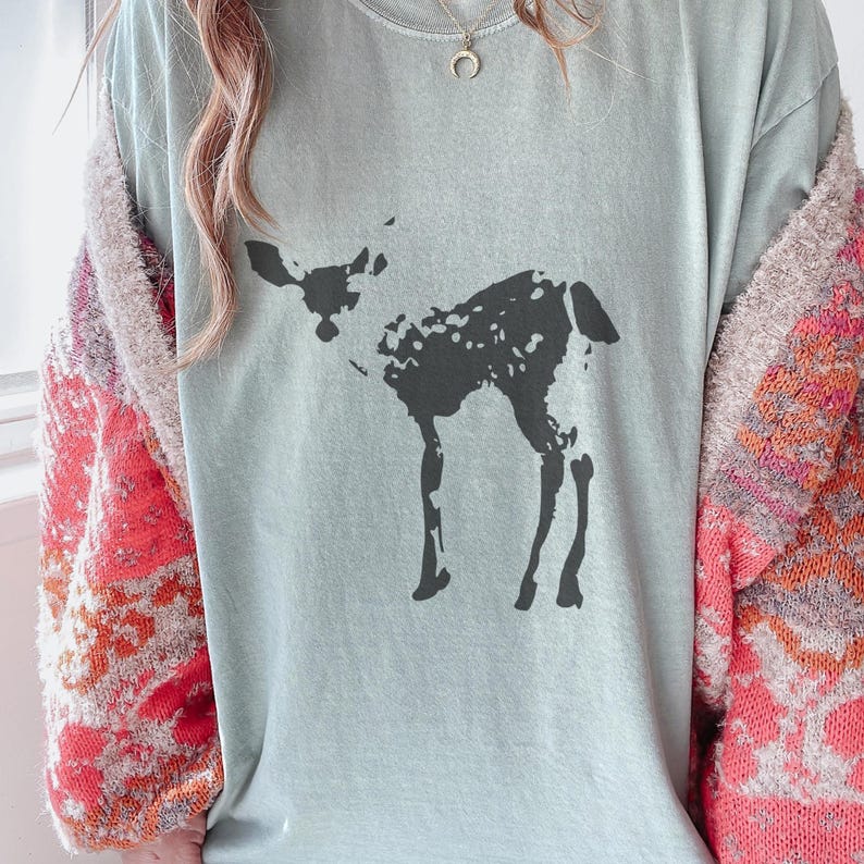 May include: A light blue t-shirt featuring a dark gray illustration of a deer. The deer is depicted in a simple, stylized manner. The shirt is worn with a patterned cardigan.