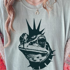 Peut inclure: Un t-shirt à manches courtes vert sauge avec une impression graphique vert foncé représentant une grenouille avec une crête iroquoise à pointes et un collier à pointes. Le t-shirt a un col rond et une coupe décontractée.