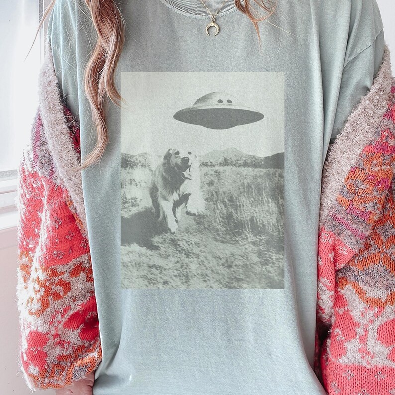 May include: A light blue t-shirt with a black and white image of a dog looking up at a flying saucer. The dog is standing in a field of grass.