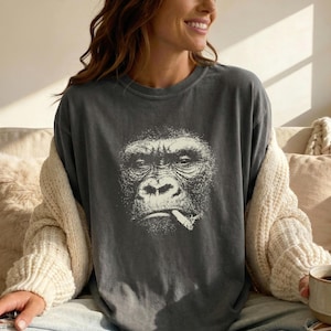May include: Dark gray t-shirt featuring a detailed, black and white illustration of a gorilla smoking a cigarette. The shirt is a relaxed fit and has a crew neck. The gorilla's face is the focal point of the design.