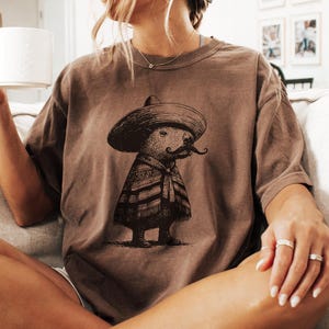 May include: Brown t-shirt featuring a whimsical illustration of a creature wearing a sombrero, poncho, and a large mustache. The graphic is in black and white. The shirt is a neutral color.