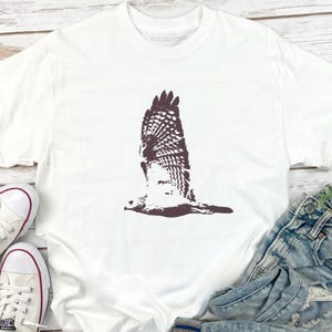 May include: White t-shirt featuring a brown graphic of a hawk in flight. The bird's wings are spread wide, showcasing detailed feather patterns. The shirt is paired with blue denim jeans and white sneakers with red accents.