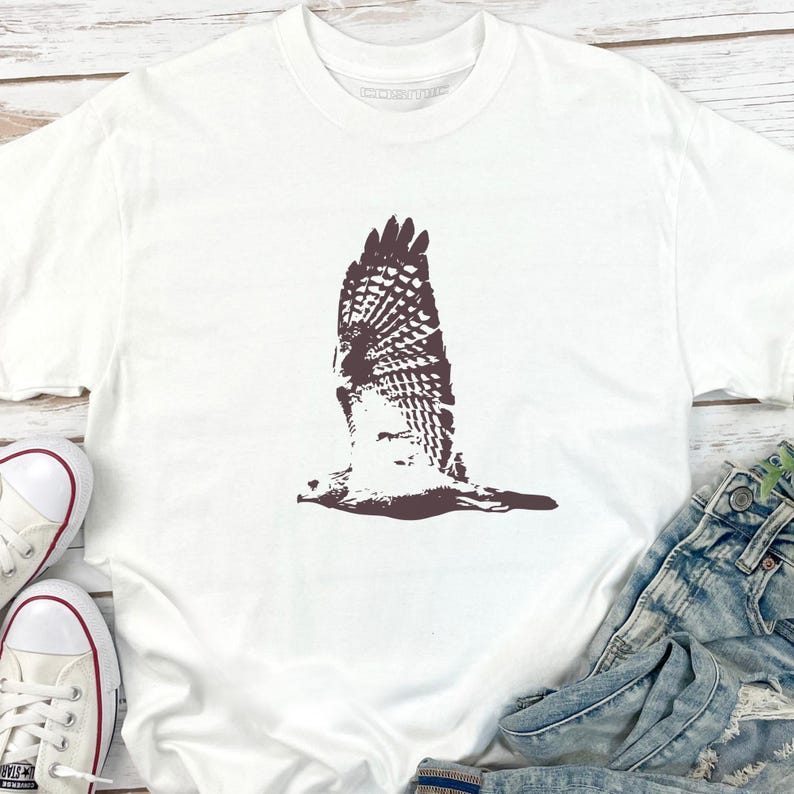 May include: White t-shirt featuring a brown graphic of a hawk in flight. The bird's wings are spread wide, showcasing detailed feather patterns. The shirt is paired with blue denim jeans and white sneakers with red accents.