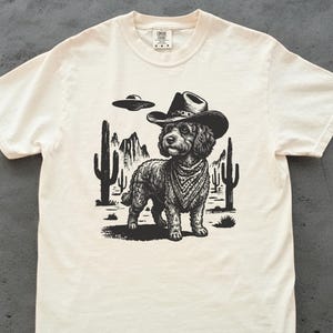 May include: A cream-colored short-sleeve t-shirt featuring a black graphic print of a dog wearing a cowboy hat and a bandana. The background of the print shows a desert landscape with cacti, mountains, and a flying saucer hovering in the sky.