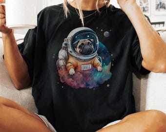 May include: A black t-shirt featuring a graphic of a pug wearing an astronaut helmet and an orange space suit. The suit has a name tag that reads CHARLIE. The pug is surrounded by a colorful nebula, stars, and planets against a dark background.