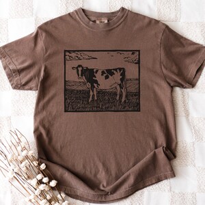 May include: Brown short-sleeve t-shirt featuring a black and white cow illustration in a rectangular frame. The cow stands in a field under a cloudy sky. The shirt has a crew neck and a relaxed fit.