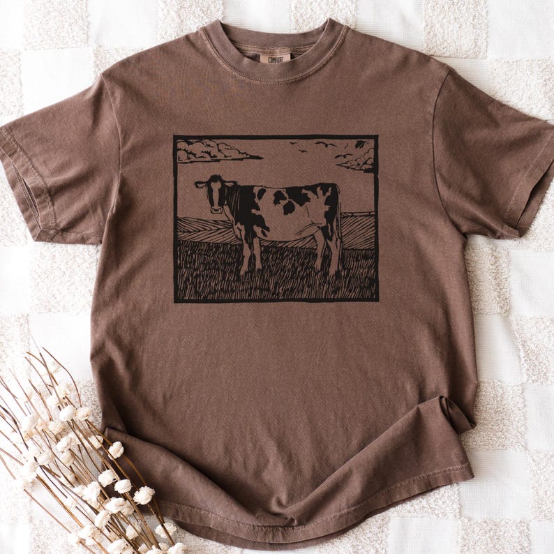 May include: Brown short-sleeve t-shirt featuring a black and white cow illustration in a rectangular frame. The cow stands in a field under a cloudy sky. The shirt has a crew neck and a relaxed fit.