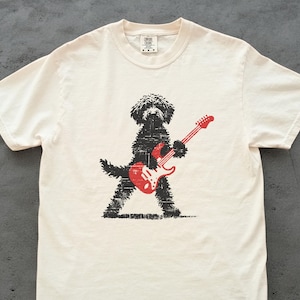 May include: Off-white t-shirt featuring a black dog playing a red electric guitar. The dog is depicted in a stylized, textured design. The guitar has red strings and a white pickguard. The shirt has a crew neck and short sleeves.
